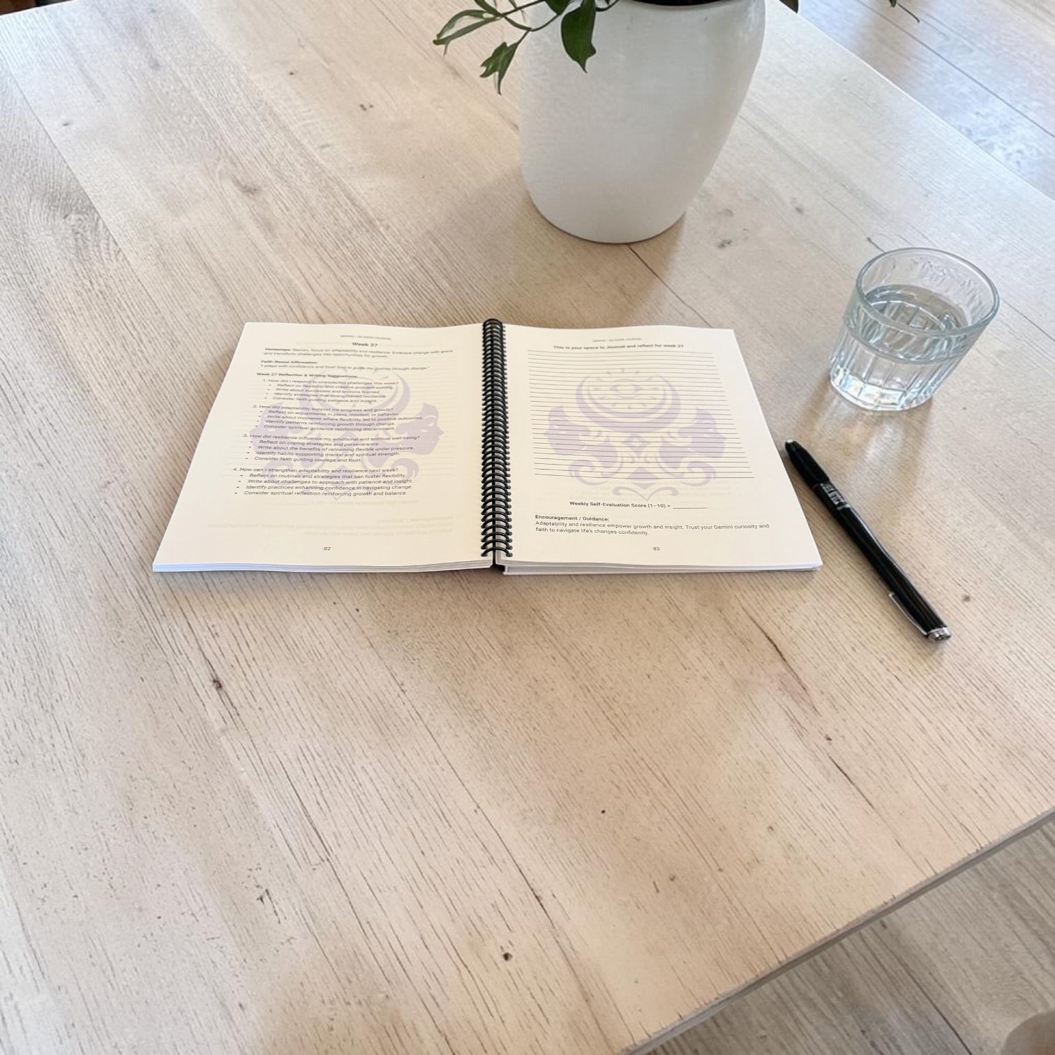 Gemini 52-Week Zodiac Journal lay-flat interior image with a pen and a glass of water on a light colored wooden table